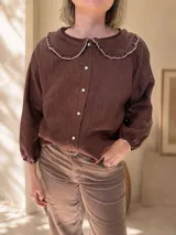 Camisa de color chocolate con textura de rayas verticales sutiles, cuello redondo con solapas redondeadas y volados, cierre frontal con botones de nácar y mangas largas con puños elásticos.