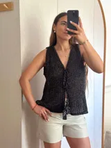 Chaleco tejido en punto calado con hilo de algodón y lurex, de color negro con destellos plateados. Presenta escote profundo en V, cierre frontal con cordones entrelazados terminados en borlas y un corte recto y holgado.