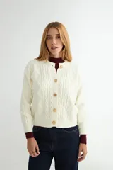 Cardigan de punto trenzado color crema con cuello redondo y cierre frontal de botones grandes en tono dorado. Presenta mangas largas con puños acanalados y un diseño de tejido con textura.