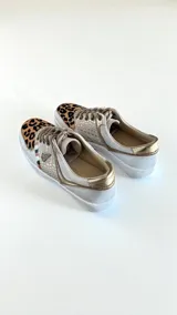 Sneakers de mujer en tonos blancos, dorados y detalles con estampado animal print de leopardo en la puntera. Incorporan cordones frontales y apliques metálicos decorativos.
