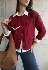 Cardigan de punto con textura trenzada en color bordó, con cuello redondo y cierre frontal mediante botones tipo alamar. Presenta mangas largas con puños acanalados y un corte clásico.