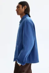 Sobrecamisa de lona de algodón en color azul, con cuello clásico, cierre frontal de botones y tres bolsillos de parche en la parte delantera. Presenta mangas largas con puños abotonados y canesú en la espalda.