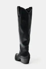 Bota alta estilo cowboy de caña alta, con tacón medio en bloque y punta fina. Presenta tiradores laterales para facilitar el calce.