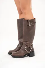 Bota biker de caña alta color marrón, confeccionada en cuero. Presenta hebilla en la parte superior, detalle de aro en la parte inferior y cierre lateral.