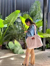 Bolso tipo tote confeccionado en corderito sintético color beige, con cierre principal y bolsillo interior. Presenta una etiqueta de cuero sintético en el frente.