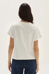 Remera de algodón color blanco, manga corta, con un estampado central que incluye una flor azul y formas abstractas con patrón a cuadros en tonos naranjas y azules, con texto bordado.