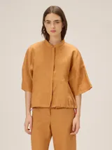 Chaqueta de lino y rayón color tostado, de corte holgado, mangas cortas amplias, cuello mao y cierre frontal oculto con cremallera. Presenta pespuntes a contratono en las costuras y cordón ajustable en el bajo.