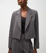 Blazer corto de sastrería color gris oscuro, con solapas estructuradas, manga larga y corte ajustado. Se combina con un top de encaje negro y pantalón ancho a juego.