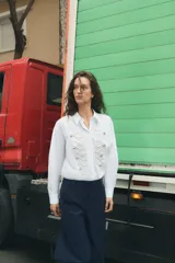 Camisa de popelín blanca con cuello solapa y manga larga. Presenta un diseño frontal con bordados en relieve y botones decorativos negros. Cierre frontal con botones ocultos.