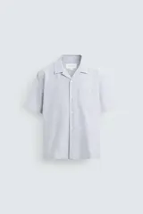 Camisa de algodón con rayas verticales finas azules y blancas, corte relajado (relaxed fit), cuello bowling y manga corta. Posee un bolsillo plastrón en el pecho y aberturas laterales en el bajo.