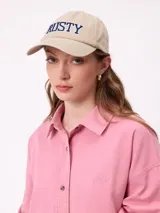 Gorro tipo gorra con visera de color taupe, con la palabra "RUSTY" bordada en relieve en color azul con contorno blanco en la parte frontal.