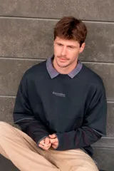 Sudadera gris oscuro de doble manga, con efecto de capas que simula una camisa a cuadros debajo, cuello redondo y bordado sutil en el pecho.
