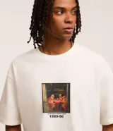 Remera de corte boxy color crudo, con cuello redondo y mangas cortas. Estampa frontal con dibujo de perros jugando a las cartas y texto 'underappreciated crew 1989-90'.