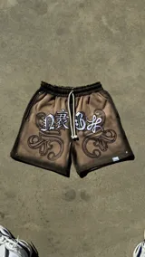 Bermuda de felpa color beige con lavado lateral negro, corte ancho. Presenta un gran estampado serigrafiado frontal con letras blancas de estilo gótico y ornamentos negros, y un aplique bordado sobre la estampa. Cuenta con cordón blanco en la cintura y remaches metálicos cromados.