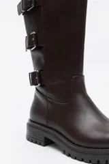 Bota de caña alta tipo biker en símil cuero color marrón oscuro. Presenta múltiples tiras horizontales con hebillas metálicas laterales y una suela robusta con relieve.