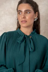 Blusa plisada de color verde oscuro, con mangas largas terminadas en puños elásticos y cuello alto con lazo frontal para anudar.