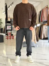 Sweater de punto liviano color beige, con textura marcada, cuello redondo y mangas largas de corte relajado, con pequeño estampado tipográfico negro en el lateral inferior.