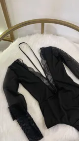 Body de lycra y encaje color negro, con mangas largas de tul transparente y detalle de encaje en los puños. Presenta un escote profundo con detalle de encaje y un collarín integrado que se conecta al centro del pecho.
