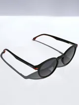 Lentes de sol con armazón redondo de color negro y detalles en naranja en las bisagras y puntas de las patillas. Cuentan con cristales oscuros polarizados y protección UV 400.