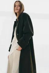 Trench oversize de color verde oscuro, con cuello de piel en tono marrón a contraste. Presenta manga larga con trabillas en los puños, bolsillos delanteros, cierre frontal con cremallera y botones ocultos bajo solapa, y abertura en la espalda.