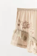 Shorts satinados color beige, con tiro medio, cintura elástica y diseño fluido. Presentan bordados florales en los laterales y volantes con puntillas en el bajo.