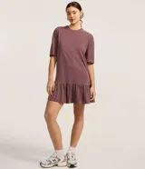 Vestido tipo camiseta de algodón, color malva o ciruela con efecto lavado, de corte recto y holgado, con cuello redondo y mangas cortas. Presenta un volado fruncido en el bajo.