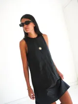 Vestido corto de gamuza sintética negra, sin mangas, de corte recto y largo asimétrico por encima de las rodillas. Cuenta con bolsillos plaqué amplios en el frente y detalle bordado de un sol en el pecho.