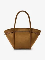 Bolso tipo shopper de eco suede color marrón, con forma trapezoidal y asas de hombro. Presenta detalles de cuero marrón con pespuntes blancos y un broche metálico dorado en el centro superior. Incluye correa larga desmontable.