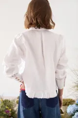 Camisa de lino blanca con cuello mao, manga larga y botones frontales. Presenta volados delanteros con terminaciones desflecadas y detalles de costuras en hilo rojo.