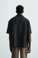 Camisa de lino color negro con calce relajado, cuello bowling y manga corta. Presenta cierre frontal con botones a tono y aberturas laterales en el bajo.