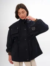 Trench azul marino con cuello alto, cierre frontal de botones a presión y cordón ajustable en la cintura. Presenta cuatro bolsillos frontales con solapa y un parche con el logo de la marca en el pecho.