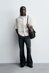 Camisa negra de corte relajado con tejido semitransparente de estructura abierta y patrón a cuadros. Presenta cuello solapa, manga corta, bolsillo plastrón en el pecho y cierre frontal de botones.