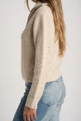 Cardigan de punto acanalado en color beige melange con motas oscuras. Presenta un diseño ajustado al cuerpo, cierre frontal completo con cremallera metálica y cuello alto.
