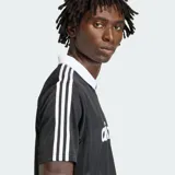 Camiseta Adidas Originals de manga corta, color negro con sutiles rayas verticales, cuello polo en V de color blanco, al igual que las líneas características de la marca en los hombros. Cuenta con el logotipo de la marca y la inscripción "adidas" en el pecho en color blanco.