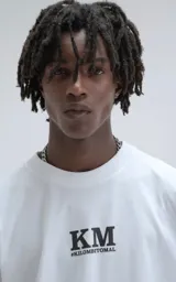 Remera blanca de corte oversize y hombros caídos, confeccionada en algodón premium. Presenta cuello redondo acanalado y estampado frontal con las siglas KM y el hashtag #KILOMBITOMAL en color negro.