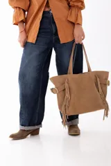 Bolso shopper de cuero descarne color tostado, con diseño de flecos laterales, hebillas metálicas decorativas y doble asa de hombro.