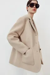 Abrigo corto de Zara confeccionado en hilatura con lana, de color beige. Presenta cuello solapa, manga larga, cierre frontal con botones negros y bolsillos de plastrón en el delantero.