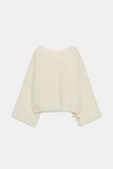 Camisa tipo capa de color beige, con cuello redondo y escote en pico en la espalda. Presenta mangas largas amplias y bajo ajustable con lazada lateral.