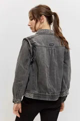 Campera de jean color gris con efecto acid wash, corte clásico y cierre frontal de botones metálicos. Presenta cuello camisero, bolsillos ribeteados y está decorada con apliques de tachas plateadas en el cuello, el canesú frontal, los bordes de la botonera, los bolsillos y el ruedo.