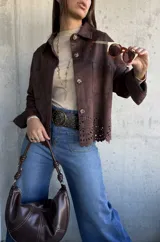 Chaqueta de gamuza color chocolate, con corte recto, cuello clásico y cierre frontal con botones. Presenta un diseño calado decorativo en el ruedo y bolsillos frontales tipo parche.