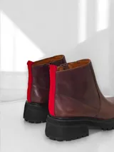 Bota corta de cuero marrón con suela track negra, cierre lateral y detalle de tira de tela roja en la parte trasera.