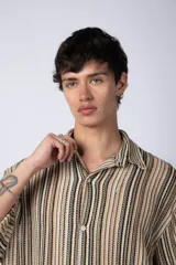 Camisa de manga corta con cuello solapa, corte holgado y cierre frontal de botones. Presenta un diseño de rayas verticales en tonos beige, negro y blanco.