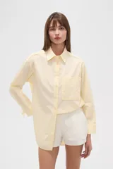 Camisa de algodón holgada, color amarillo claro con rayas finas blancas. Presenta cuello camisero, cierre frontal con botones, un bolsillo de parche en el pecho y mangas largas con puños ajustables con cordón.
