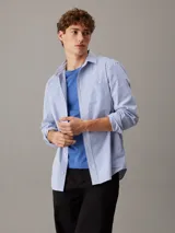 Camisa de manga larga Calvin Klein confeccionada en popelina de algodón elástico con diseño de rayas verticales. Presenta cuello en punta, cierre frontal de botones, bolsillo de parche en el pecho con logo bordado y bajo redondeado.