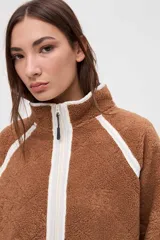 Campera de sherpa color marrón con diseño capitoneado, cierre frontal completo y detalles en color blanco. Presenta cuello alto, bolsillos laterales y puños elásticos.