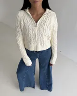 Sweater tipo campera de tejido de punto con diseño de trenzas, color blanco, con cuello alto y cierre frontal completo.