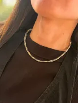 Gargantilla de cuentas tejidas con patrón de camuflaje en tonos azul, blanco y cobre. Cuenta con cierre de mosquetón metálico dorado y una pequeña placa con logo grabado.
