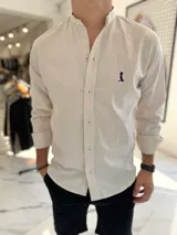 Camisa de lino color beige con cuello mao, manga larga y cierre frontal de botones. Presenta un pequeño bordado oscuro en el pecho.