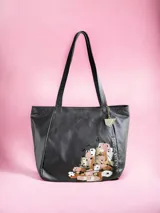 Cartera de cuero color negro, de diseño amplio con asas largas para llevar al hombro. Presenta un detalle de pintura abstracta en tonos nude y marrones en la parte inferior, y un charm metálico colgante.