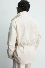 Cazadora técnica de corte relaxed fit, color beige, con cuello subido, manga larga y acolchado interior. Cuenta con múltiples bolsillos frontales con solapa, cintura ajustable con cordón interior y cierre frontal de cremallera oculta por solapa con botones a presión.